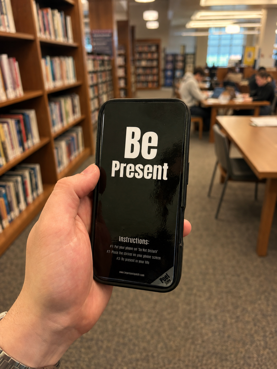 A hand holding a phone in a library with the Be Present Patch covering the screen